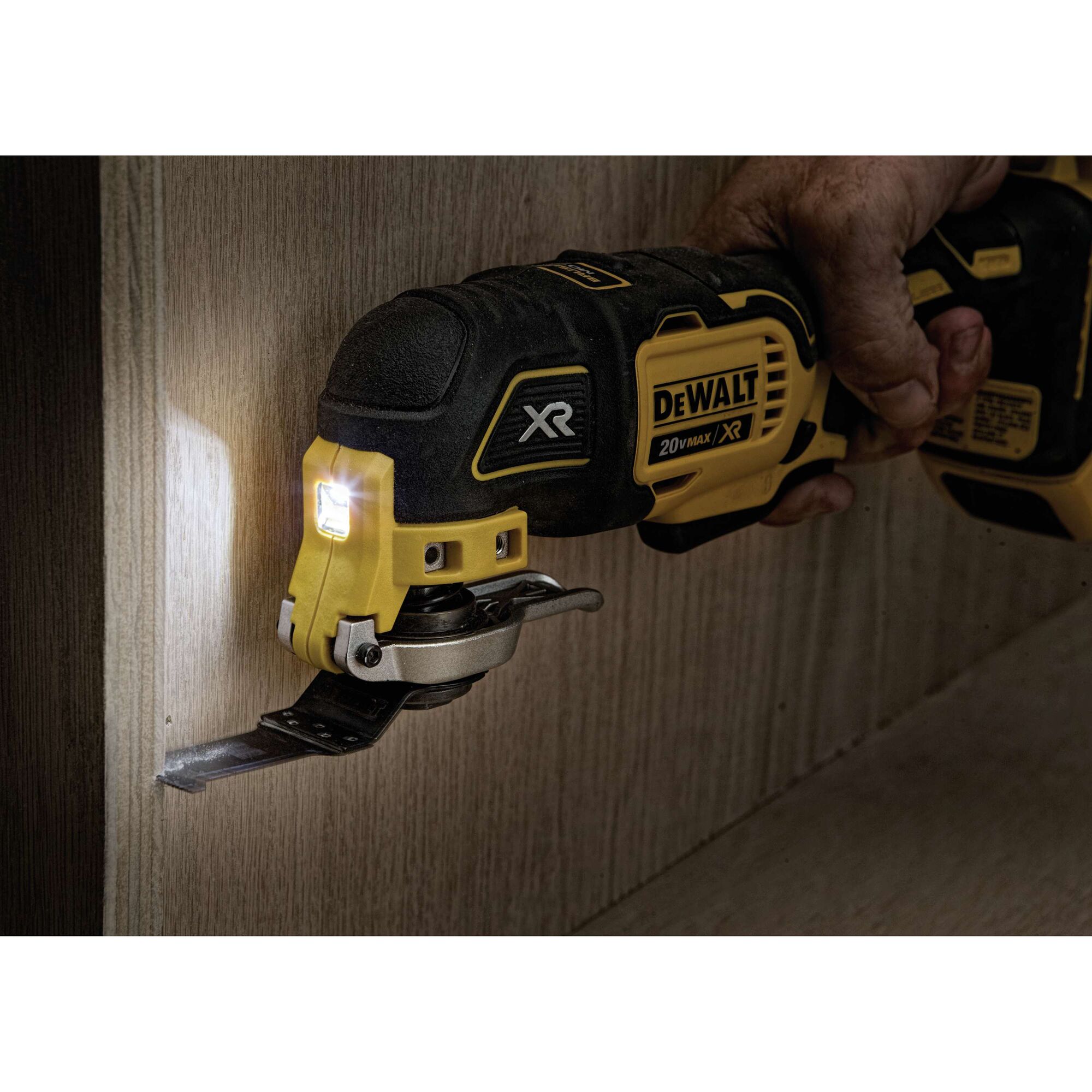 A DEWALT 20V Max XR oscillating multi-tool with a cutting blade is being used to cut into a wooden surface. The tool is equipped with an LED light, and a person's hand is gripping the handle.