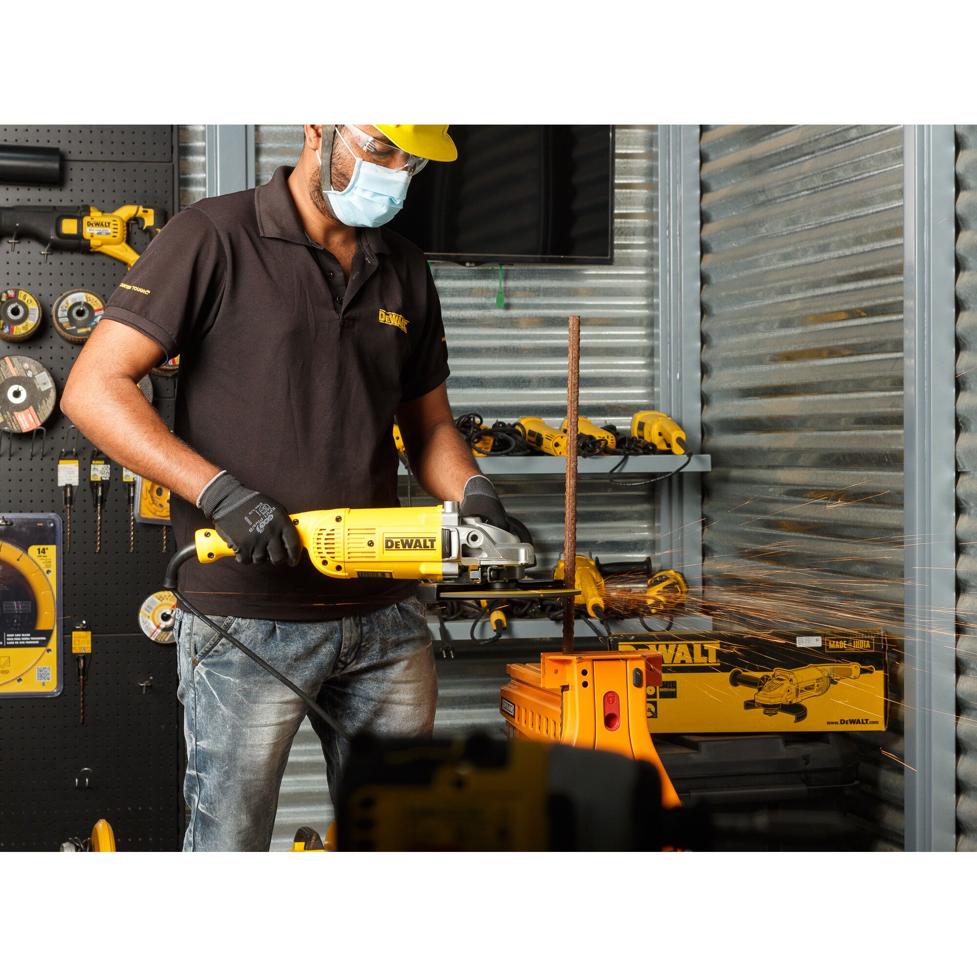 A person wearing gloves and a black DEWALT polo shirt is using a yellow DEWALT angle grinder (model DWE492) to cut a metal rod in a workshop. Sparks are visible as the tool operates. The background features various DEWALT tools and accessories organized on shelves and walls.