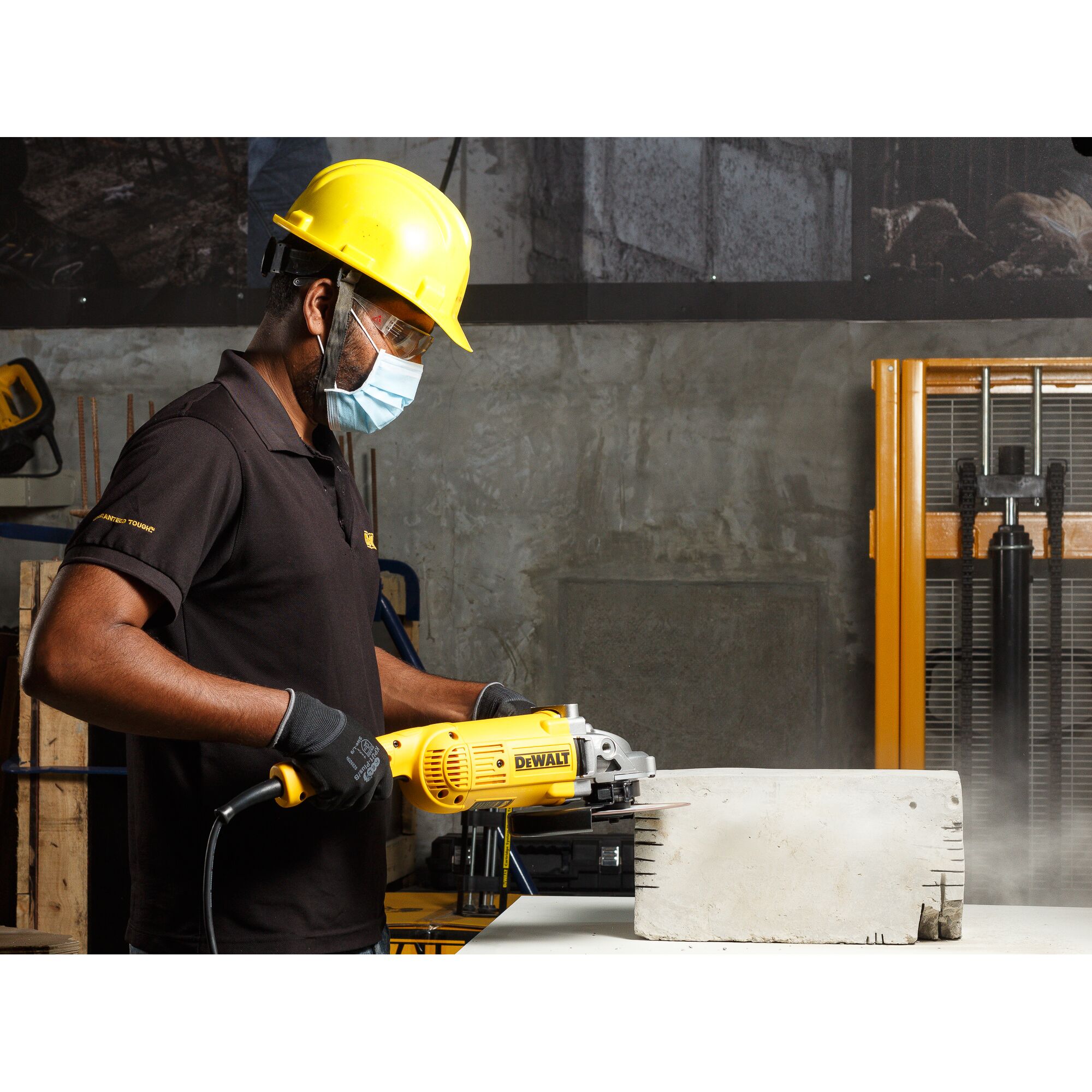 A person wearing safety gear, including a yellow hard hat and gloves, operating a DEWALT DWE492 angle grinder to cut a large concrete block in an industrial setting.