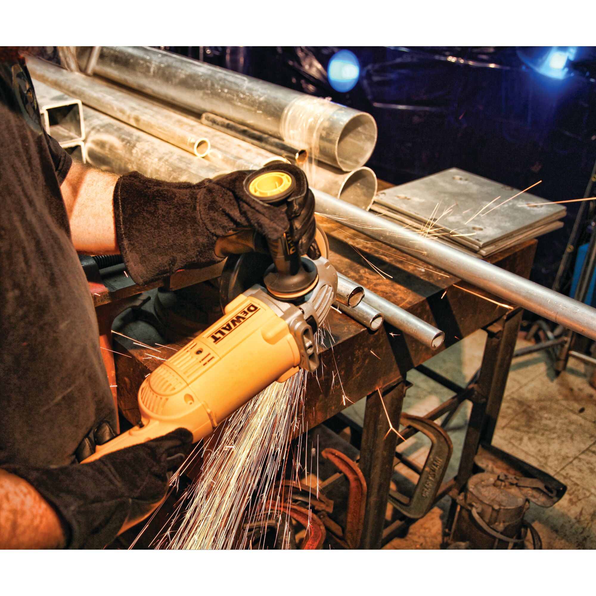 A person wearing protective gloves is using a DEWALT angle grinder to cut a metal pipe on a workbench, producing sparks. Various metal pipes and tools are visible on the workbench.