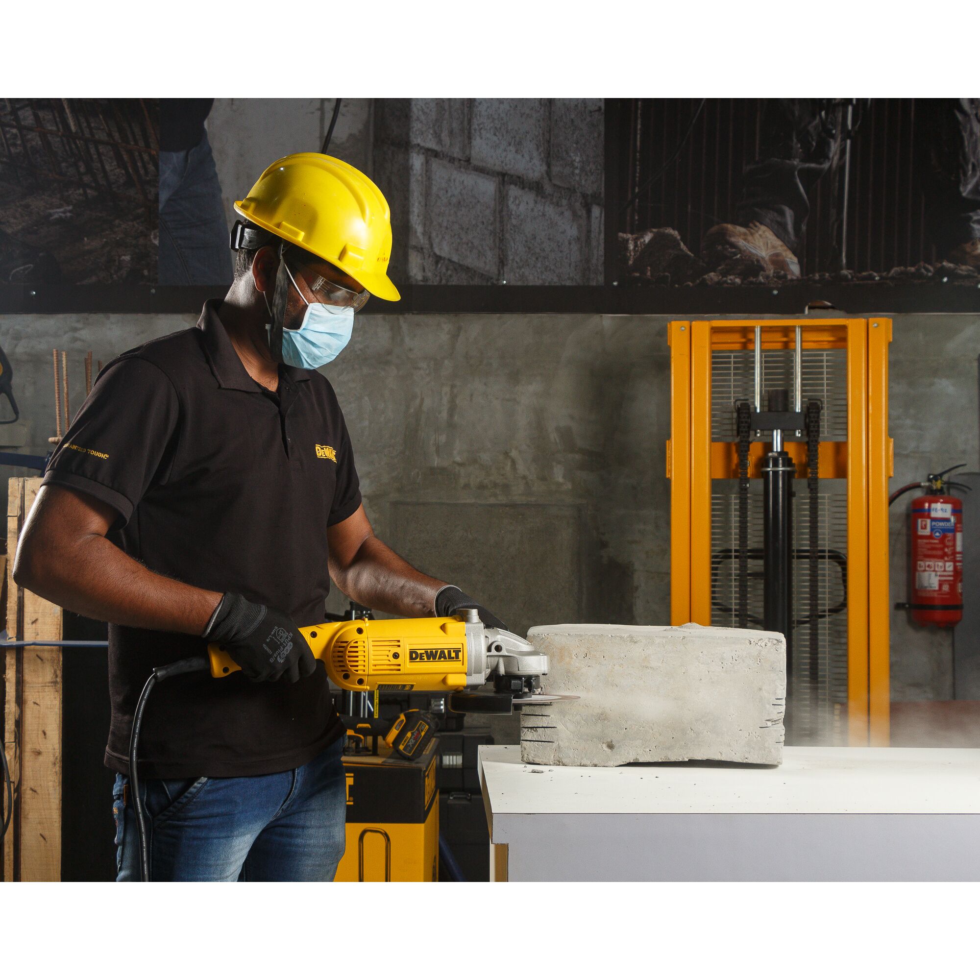 A person wearing a yellow safety helmet and black gloves is using a DEWALT DWE497 power tool to grind or cut a concrete block on a white table. The scene is set in an industrial environment with safety equipment and machinery in the background.