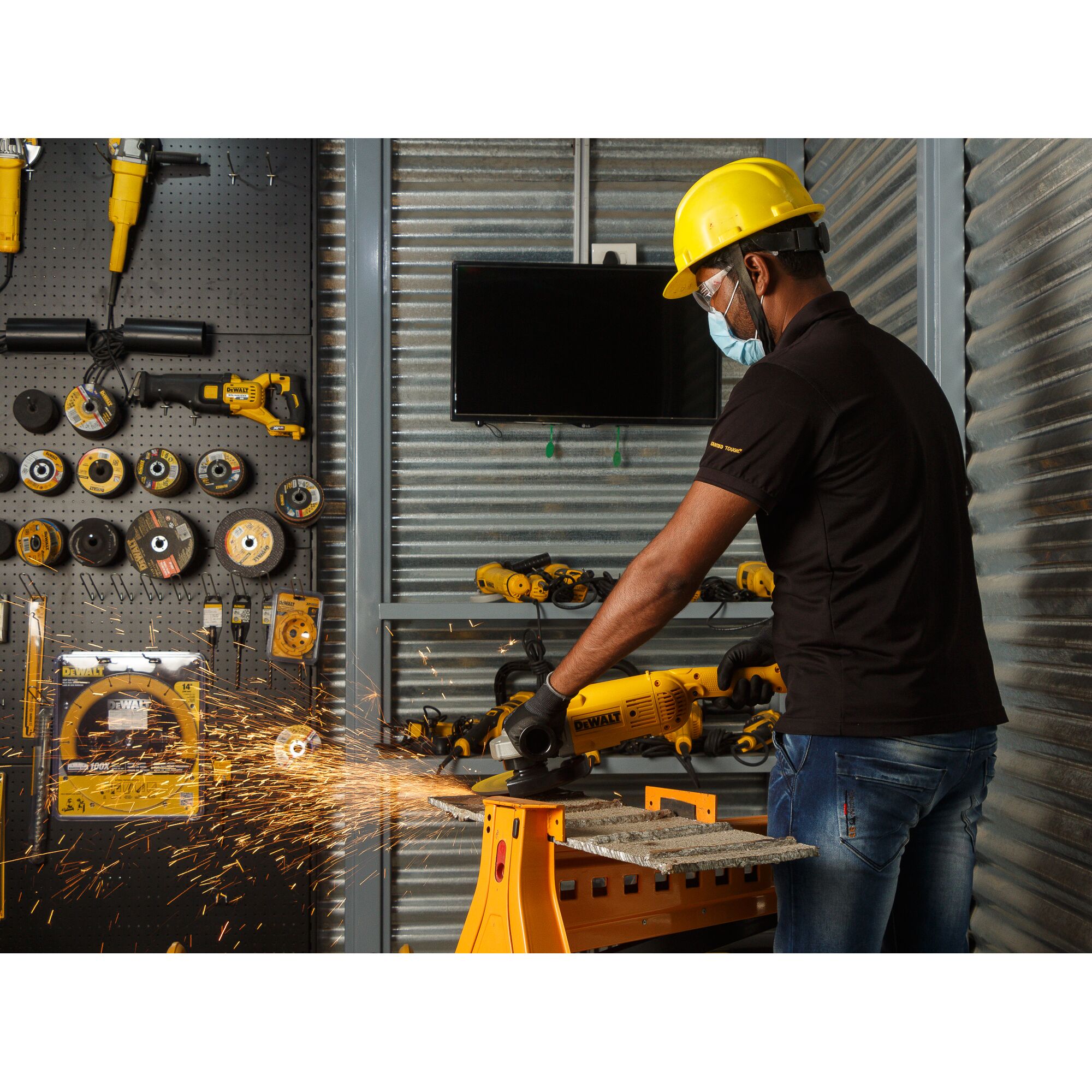 A person wearing a yellow hard hat and black gloves is using a DEWALT DWE497 angle grinder on a piece of metal, generating sparks. The scene takes place in a workshop with various DEWALT tools and equipment visible on the wall and shelves.