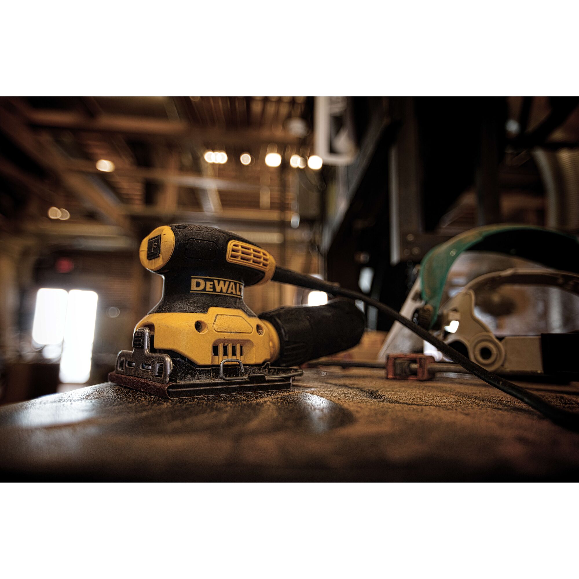 A DEWALT DWE6411 electric sander is shown on a dusty workbench in a workshop, with a power cord attached and protective gear in the background.