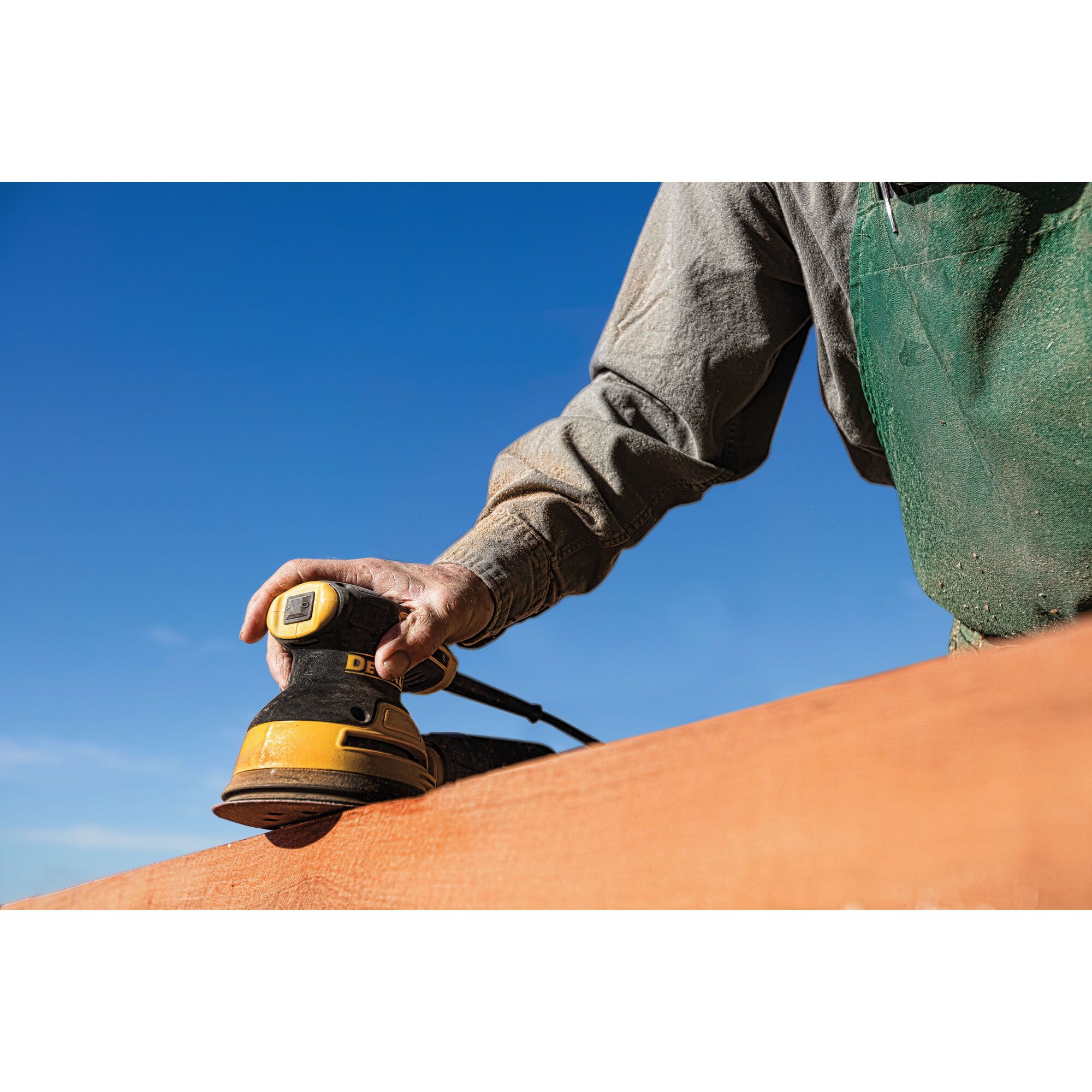 A person wearing a green apron uses a DEWALT electric sander to smooth a wooden surface outdoors under a clear blue sky.
