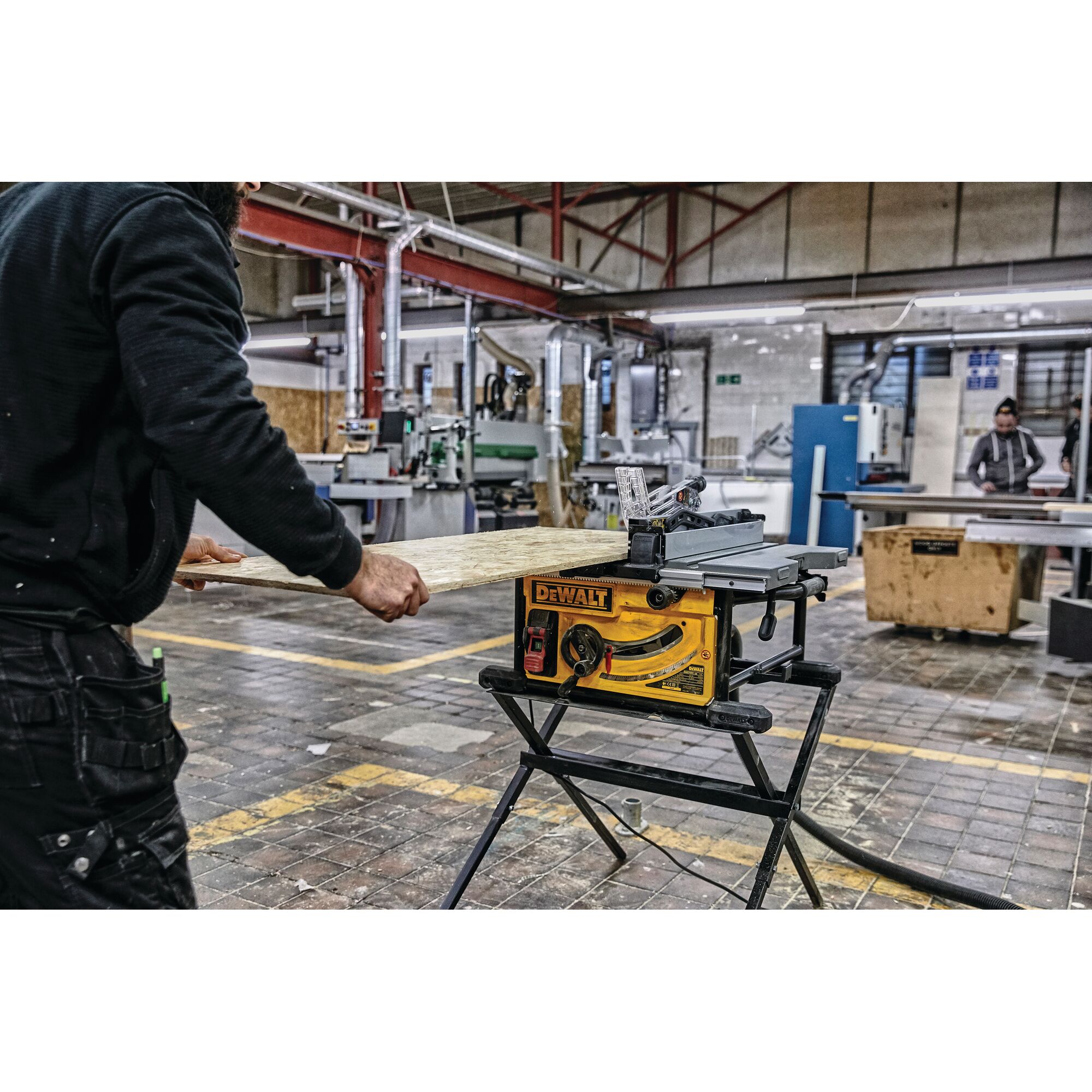 A person is using a yellow DEWALT table saw (model DWE7492SET) mounted on a black stand to cut a wooden board inside a workshop with industrial machines in the background.