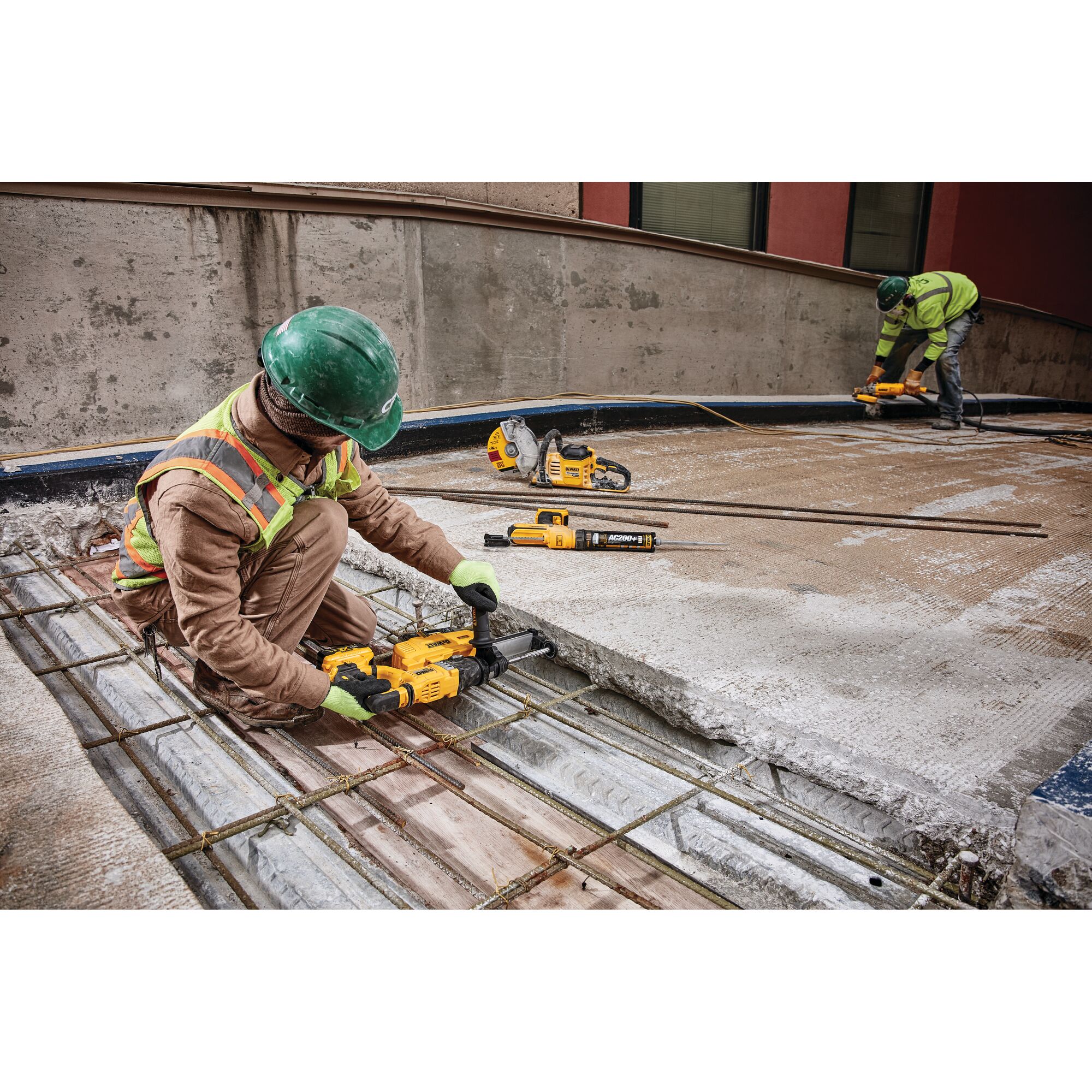 Two construction workers wearing safety gear use DEWALT power tools to cut and work with concrete and reinforcing bars on a construction site. One worker is kneeling close to the camera, using a yellow DEWALT tool, while the other is working in the background.
