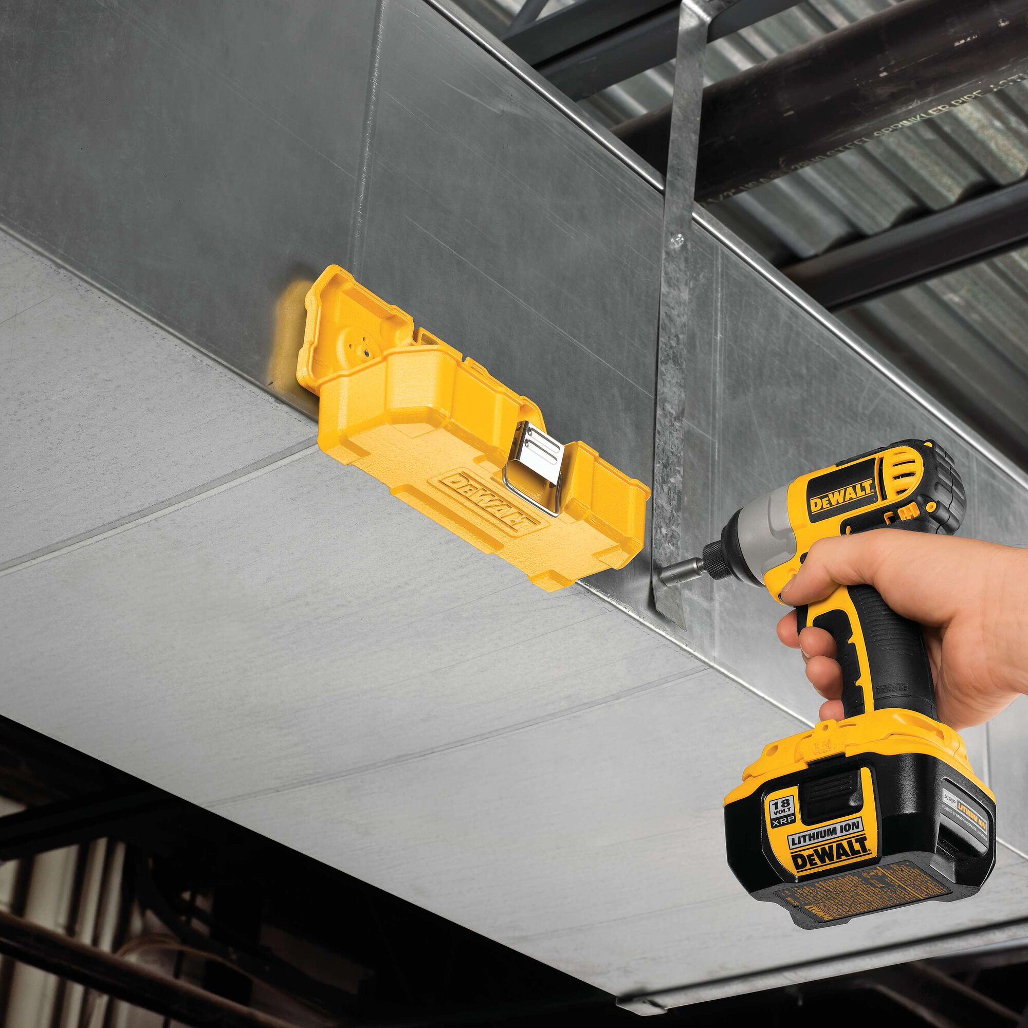 A person uses a yellow DEWALT cordless drill to attach a yellow DEWALT tool case to a metal duct or surface. The setting appears to be an industrial or construction environment.