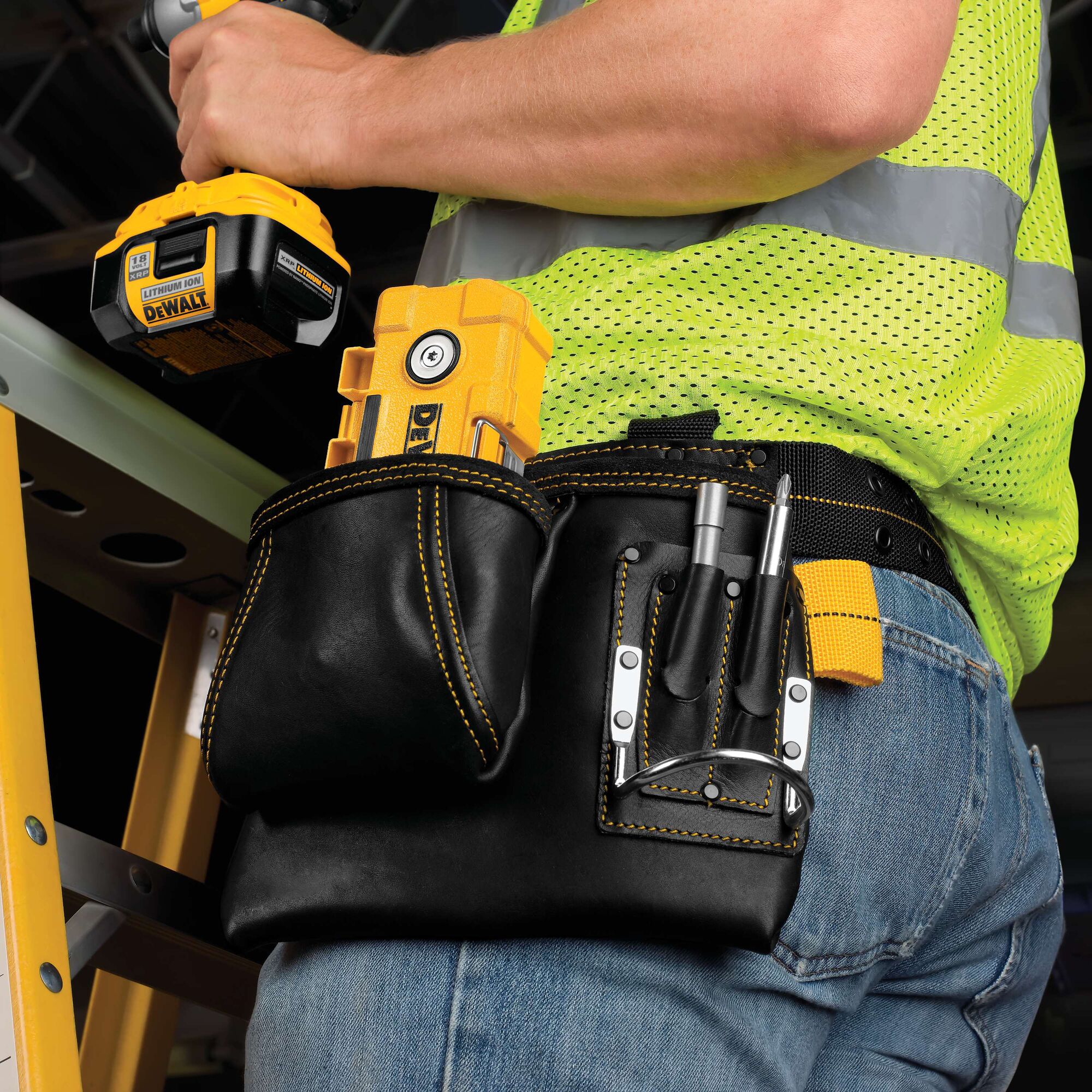 A close-up view of a person wearing a black leather tool belt with yellow stitching, containing various DEWALT tools and accessories. The person is also holding a DEWALT cordless power tool and is dressed in blue jeans and a bright yellow safety vest. A yellow ladder is visible in the background.