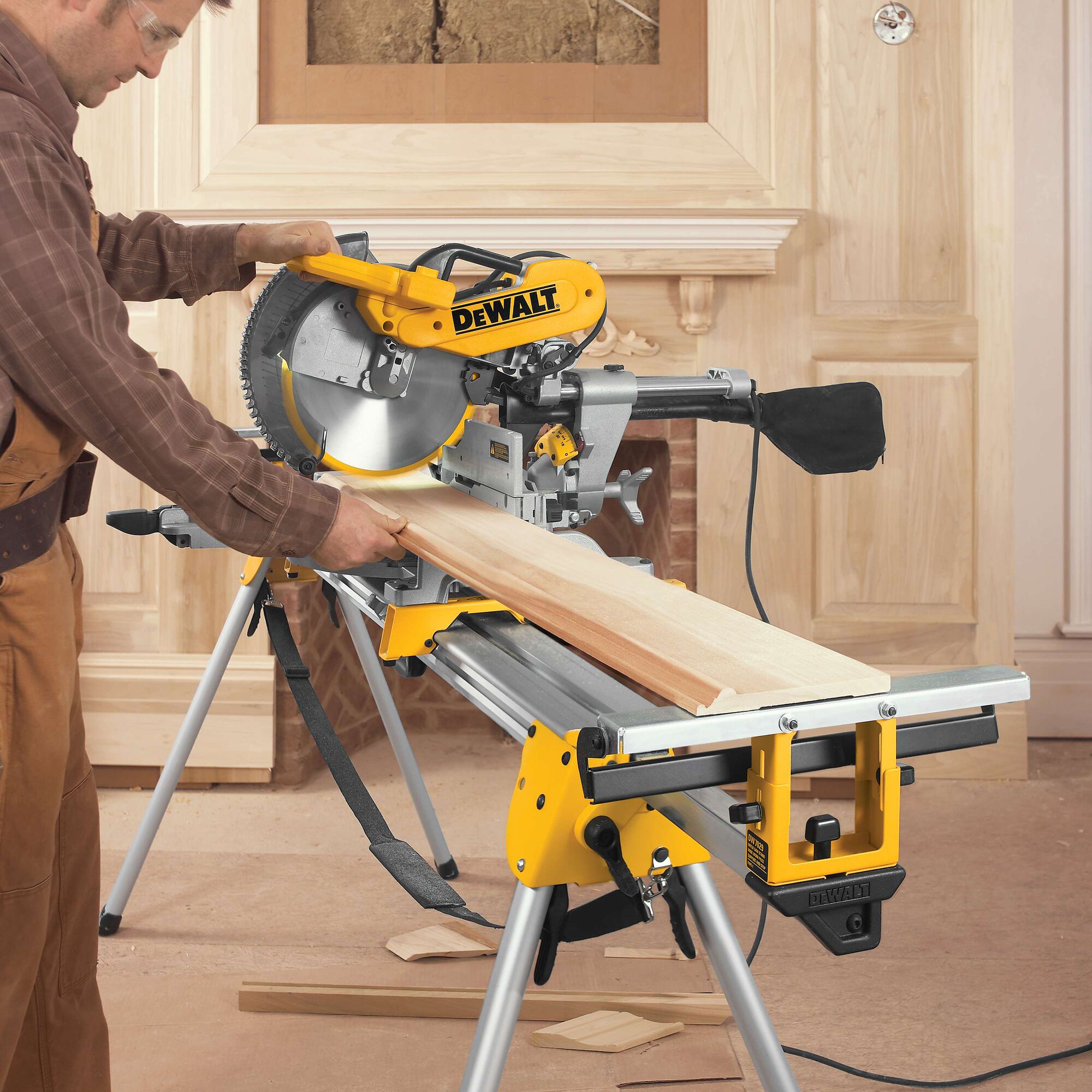 A person is using a DEWALT DWS780 miter saw to cut a wooden board in a workshop or construction setting. The saw is mounted on a stand and surrounded by wood pieces and tools.