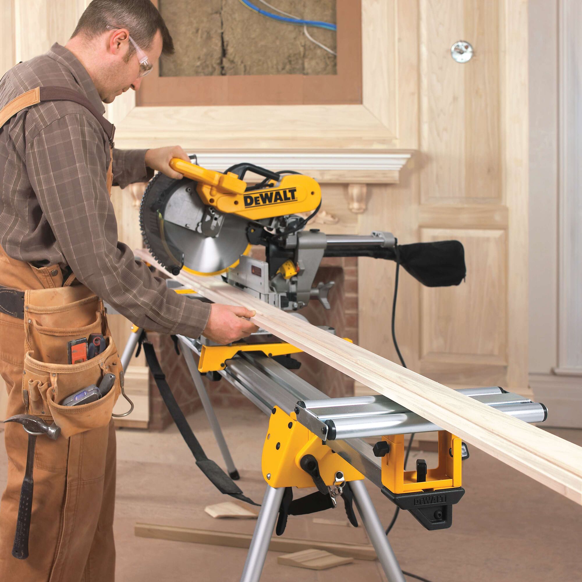 A person using a DEWALT DWS780 miter saw mounted on a stand to cut a long piece of wood. The individual is wearing a tool belt and is working in a workshop with unfinished wood panels in the background.