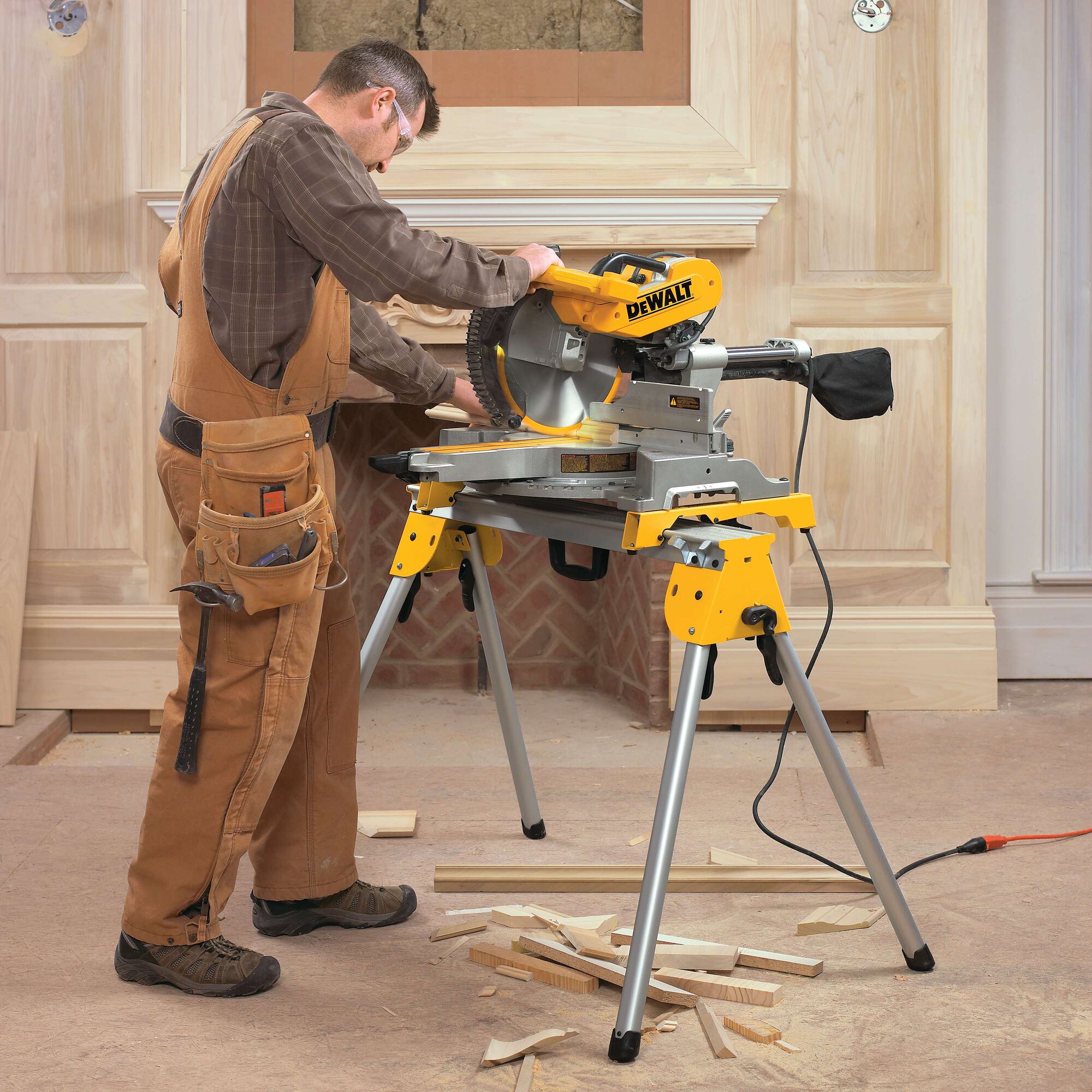 A person wearing brown overalls operates a DEWALT DWS780 miter saw on a stand indoors, cutting wooden planks. There are wood shavings and cut pieces on the floor around the saw. The workspace appears to be a woodworking or carpentry area.