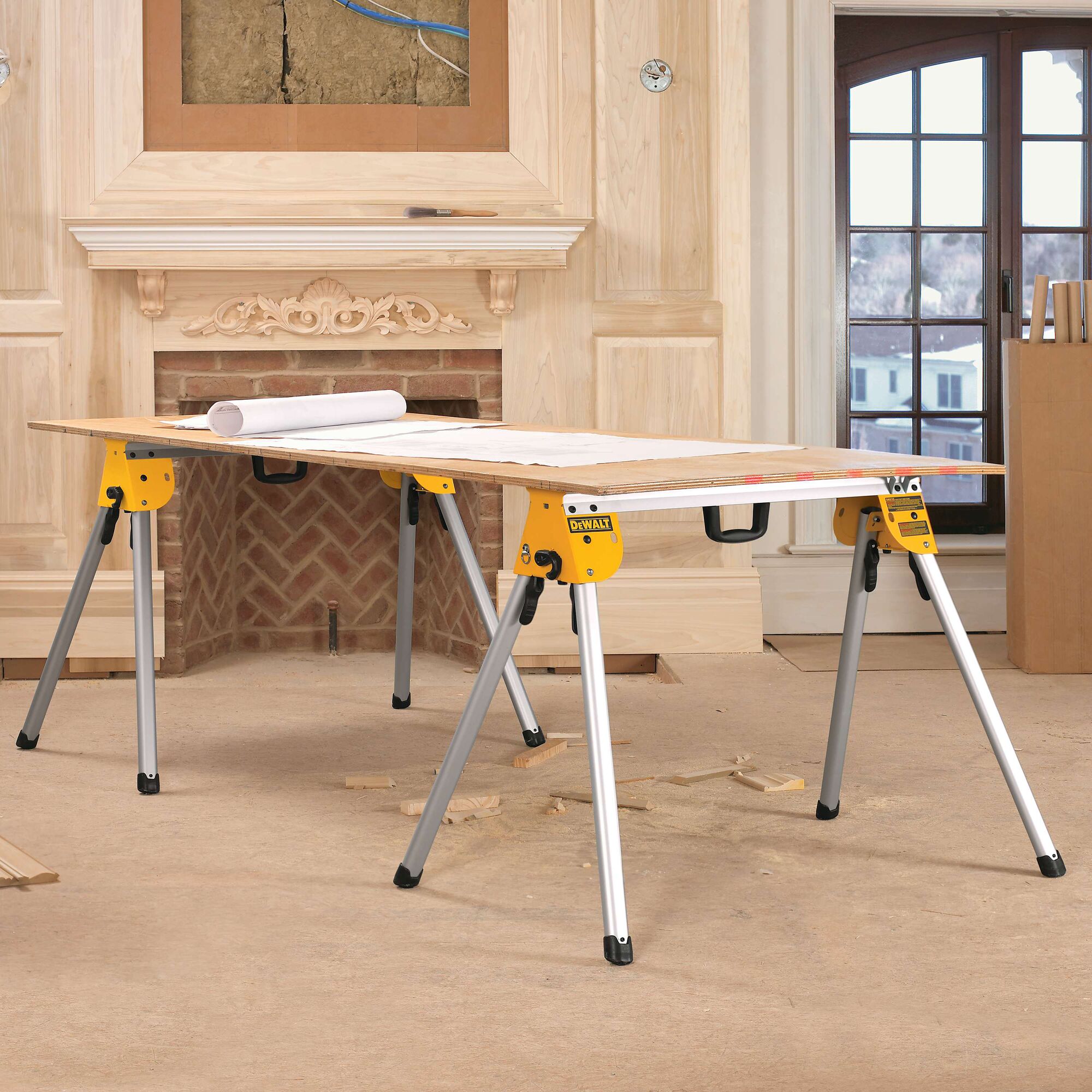 A DEWALT DWS780 yellow and silver sawhorse set used as a work table, with a wooden board and rolled plans on top, placed in a room under renovation with unfinished woodwork and a fireplace.