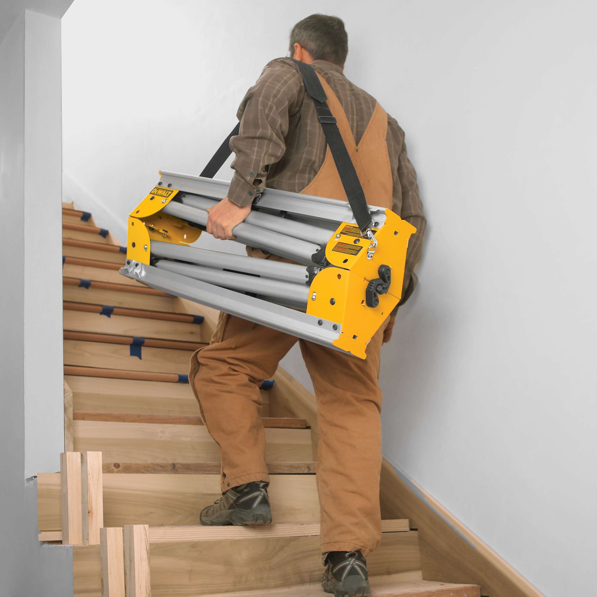 A person wearing brown work overalls is carrying a folded DEWALT tool stand with a shoulder strap while walking up wooden stairs.
