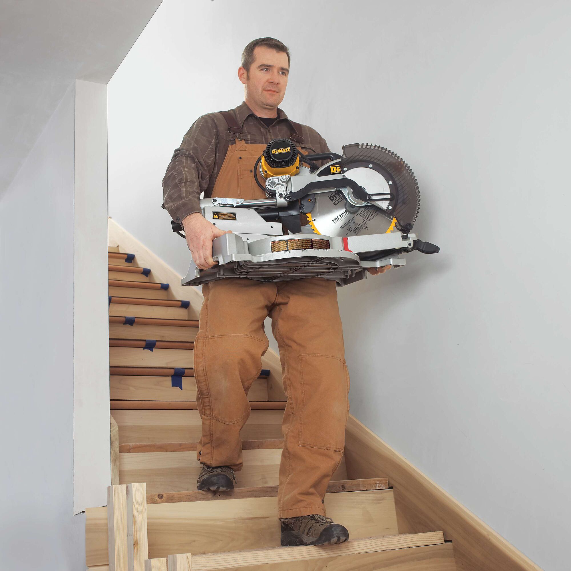 A person in brown work overalls is carrying a DEWALT DWS780 miter saw up a staircase inside a building. The face of the person is blurred for privacy.