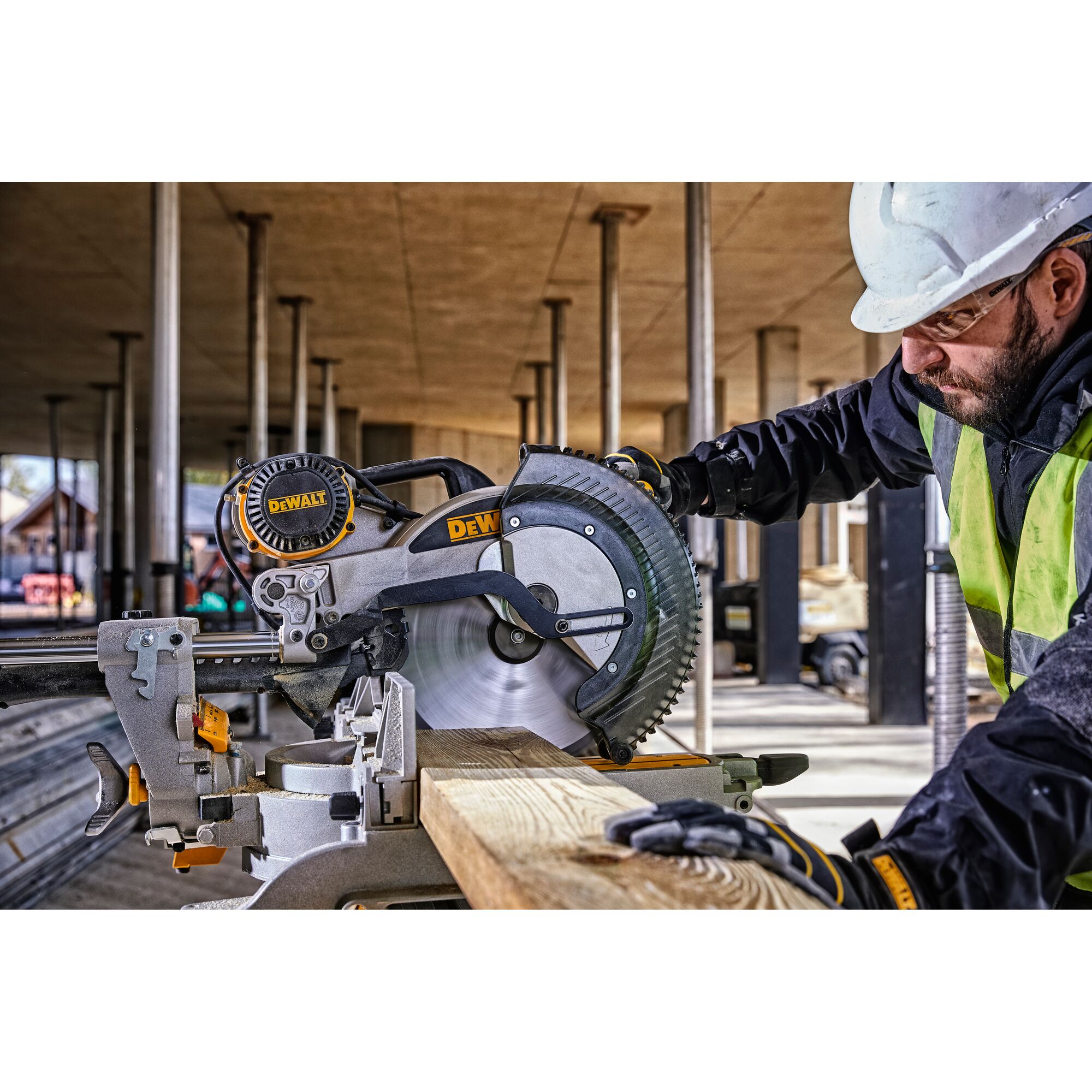 A DEWALT DWS780 miter saw is being operated by a worker wearing safety gear and a helmet at a construction site. The saw is cutting a piece of wood.