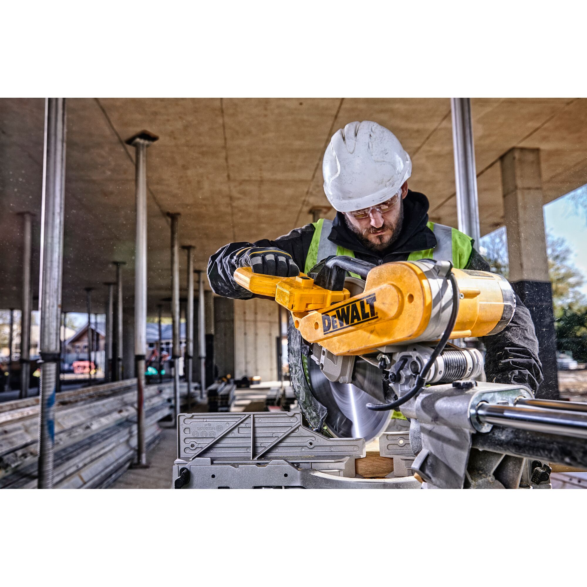 A person wearing a hard hat and safety vest is operating a DEWALT DWS780 miter saw at a construction site with metal supports and building materials visible in the background.