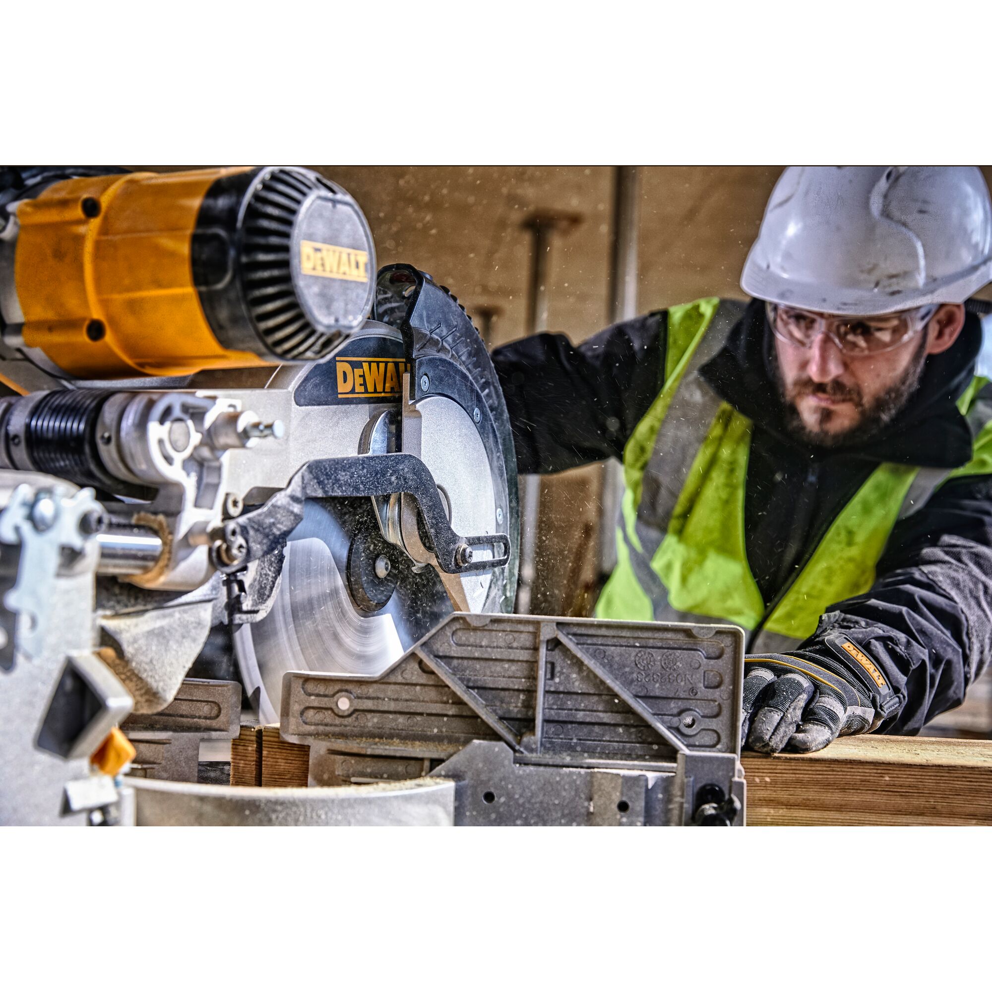 A DEWALT DWS780 miter saw being used by a person wearing a safety vest, gloves, and a hard hat. The saw is cutting through a piece of wood at a construction site.