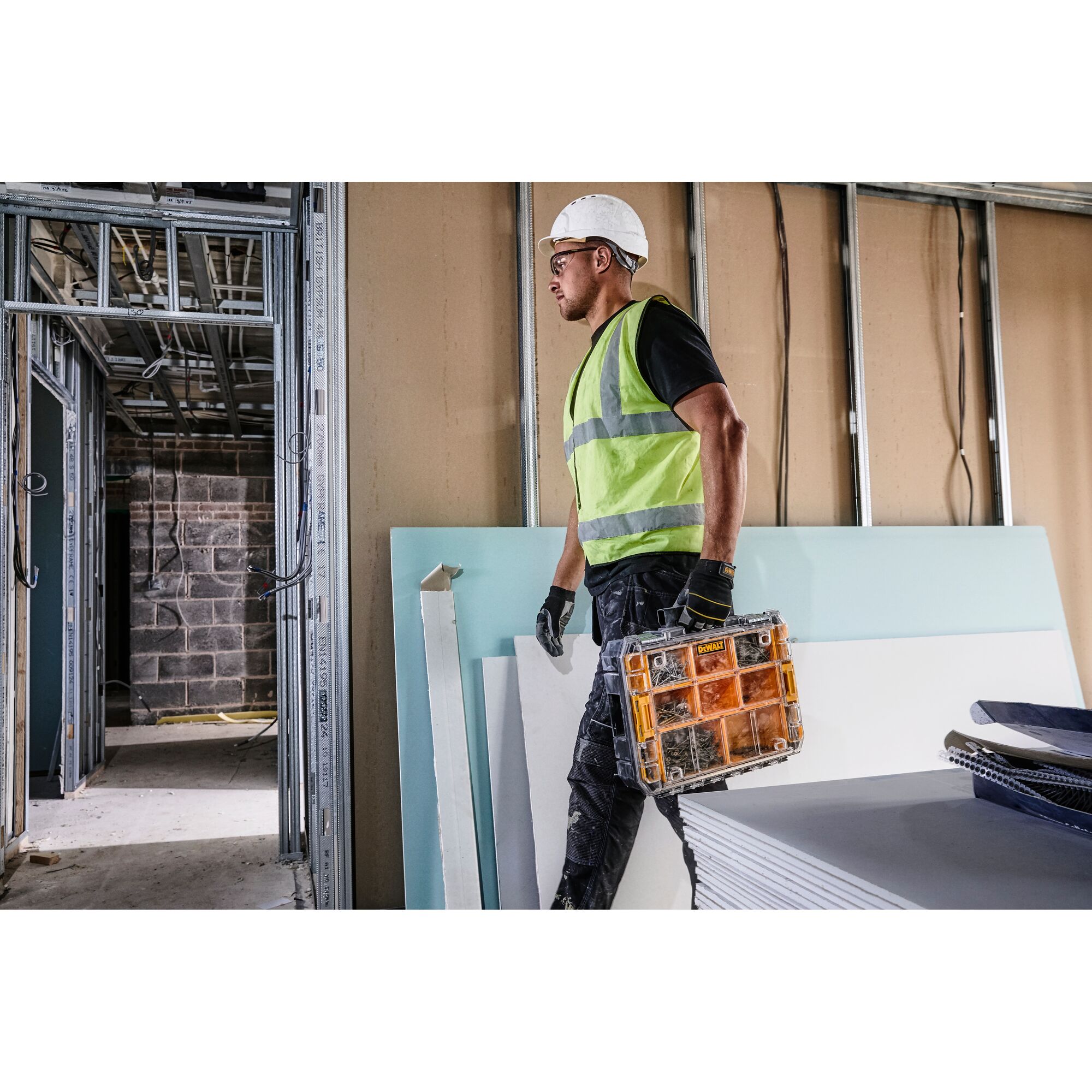 A construction worker wearing a reflective vest and helmet carries a DEWALT DWST82968-1 organizer in a building under construction.