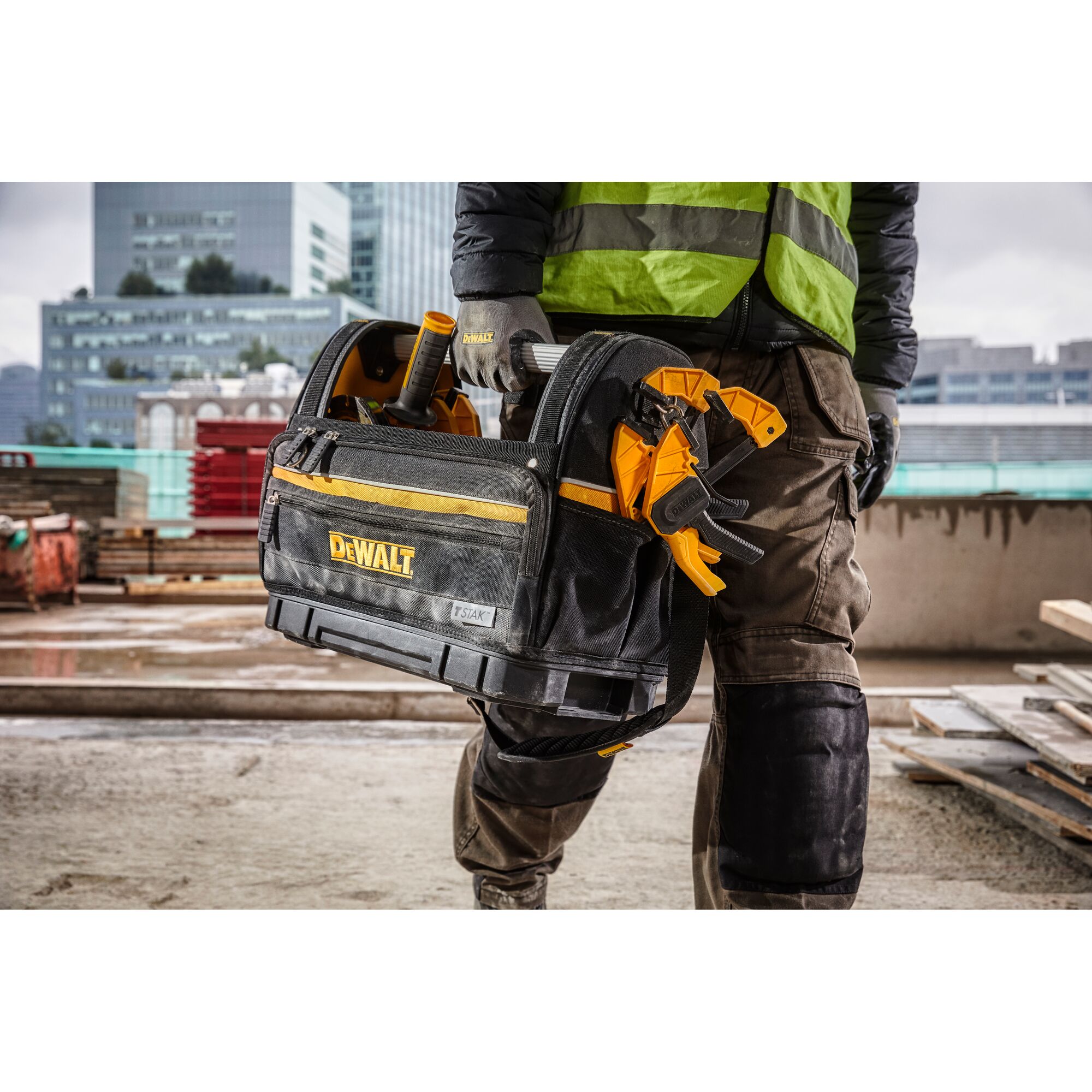 A person wearing work clothes and gloves is holding a DEWALT branded tool bag filled with various tools on a construction site. The tool bag has yellow accents and the DEWALT logo, with city buildings visible in the background.