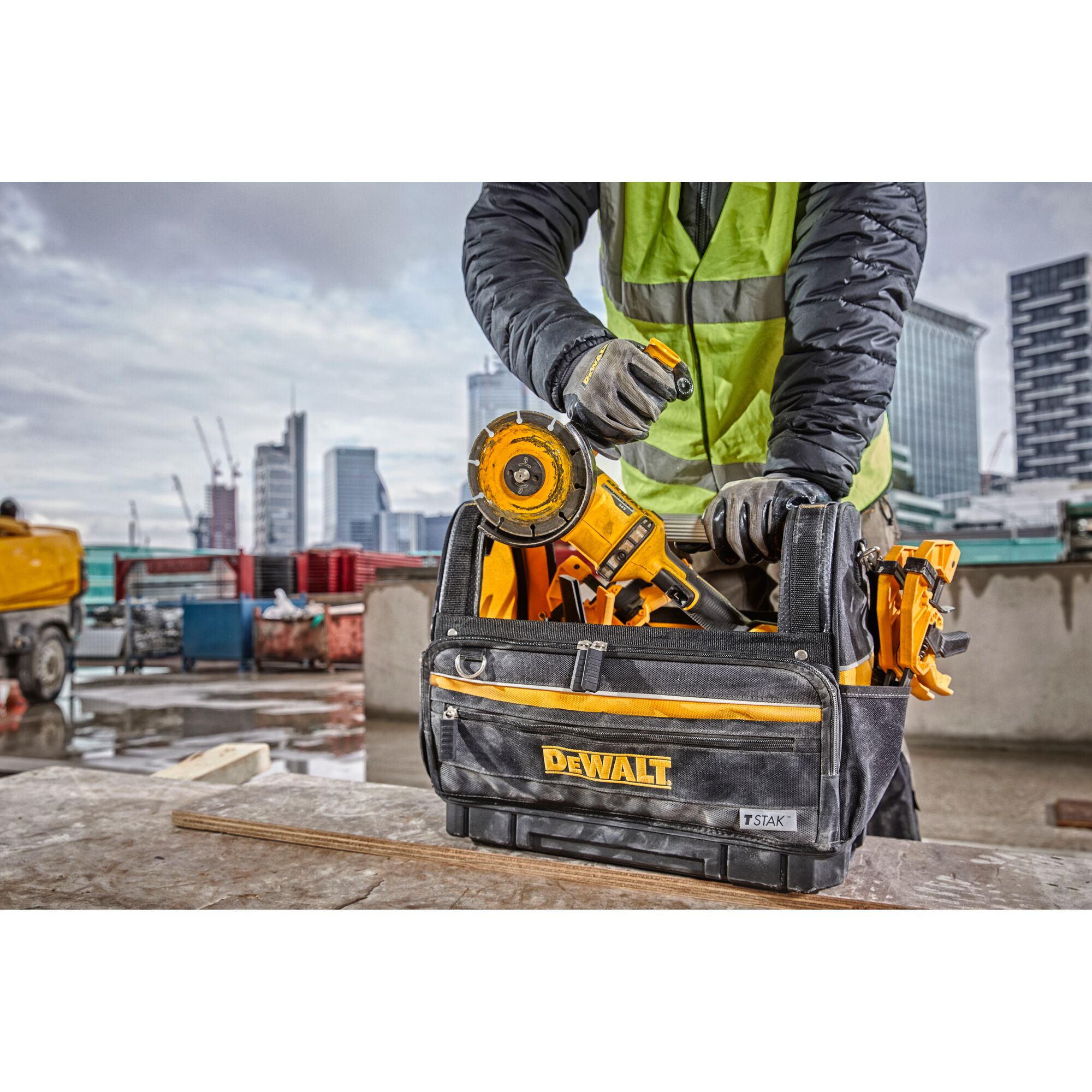 A construction worker wearing a high-visibility vest and gloves is organizing tools in a DEWALT branded tool bag, including a yellow power tool. The tool bag is placed on a work surface with an urban construction site in the background.