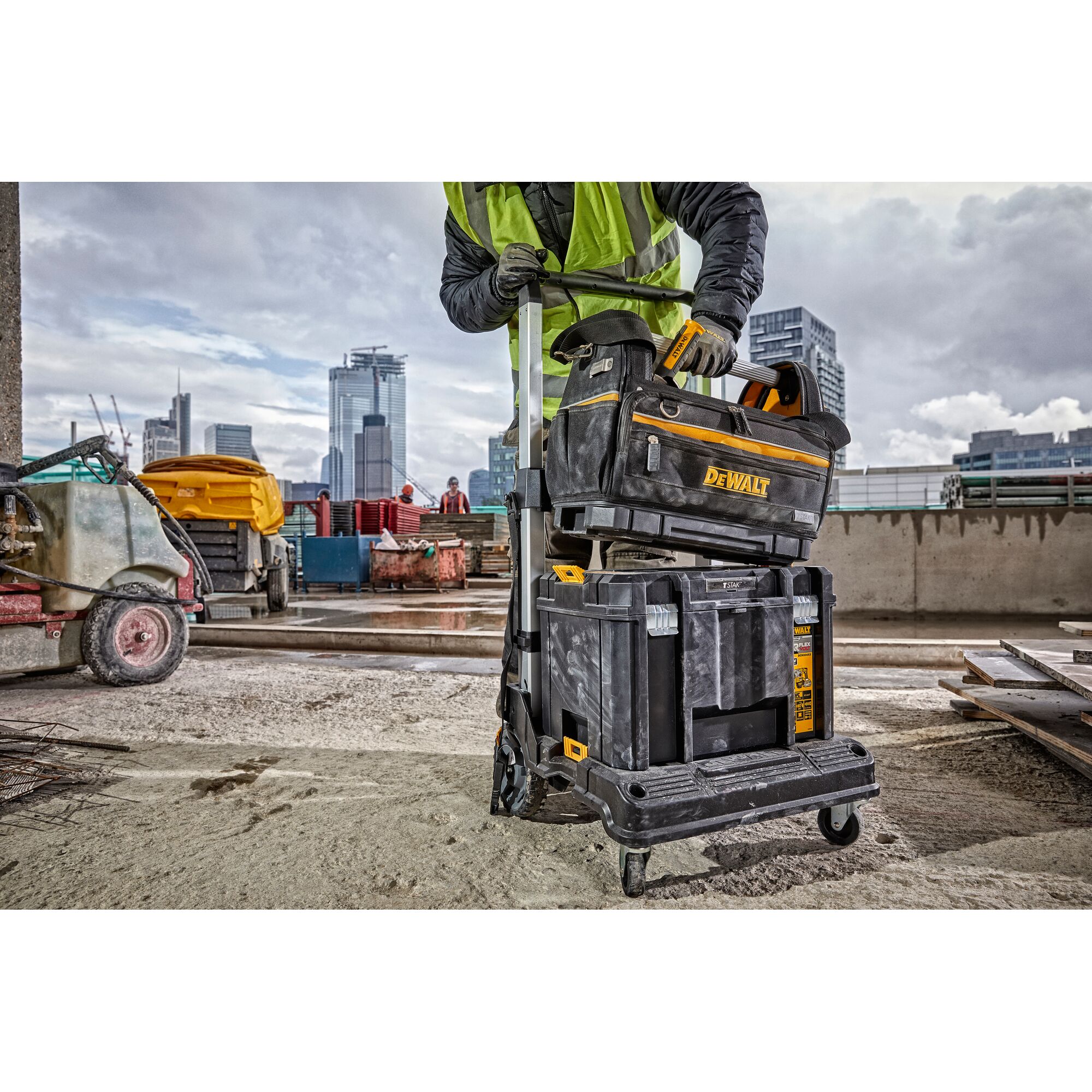 A person wearing a safety vest handles a stack of DEWALT toolboxes and a tool bag on a wheeled cart at a construction site, with modern city buildings visible in the background.