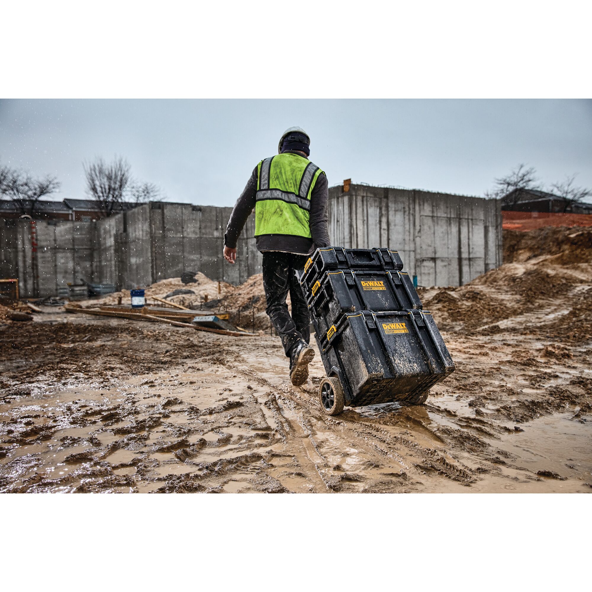 Worker pulls DEWALT toolboxes on wheels across a muddy construction site while wearing a safety vest and helmet.