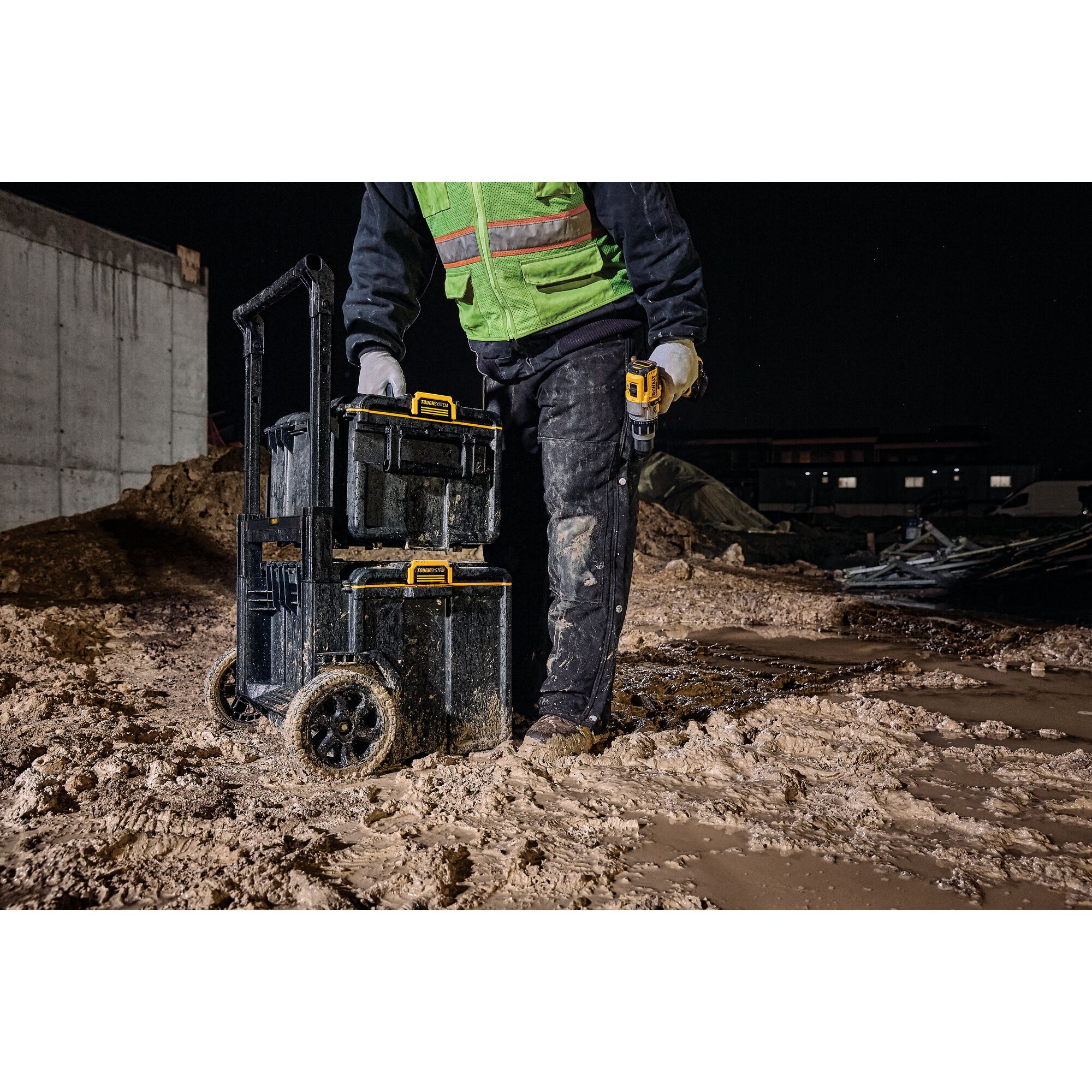 Worker with DEWALT tool boxes on wheels in muddy construction site at night.