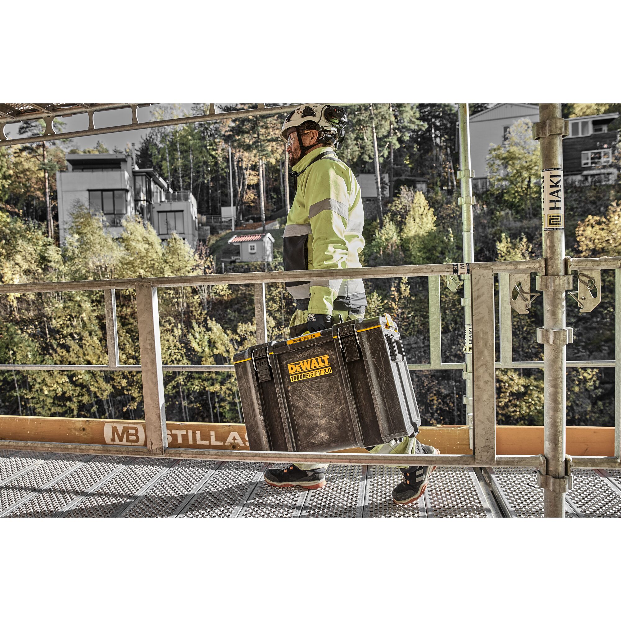 Person in safety gear carrying a DEWALT Toughsystem 2.0 toolbox on a construction site walkway.